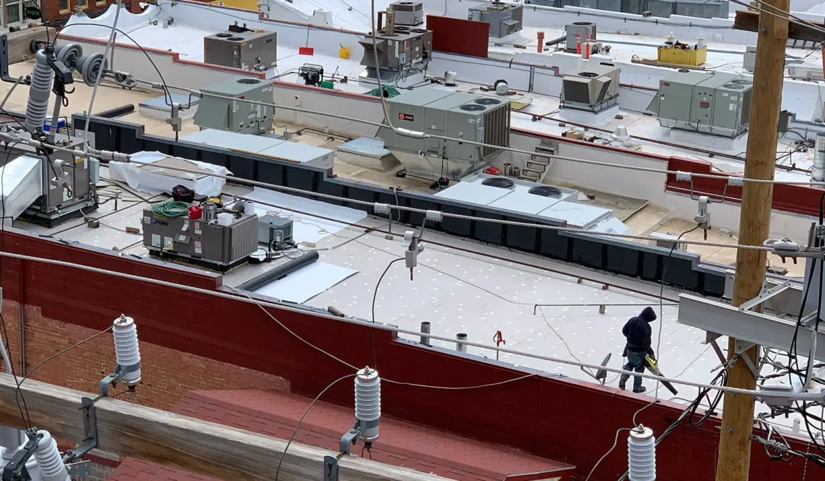 The Keys to Maintaining a Commercial Roof. Detailed view of a commercial rooftop with HVAC units, electrical wiring, and a worker conducting maintenance tasks.