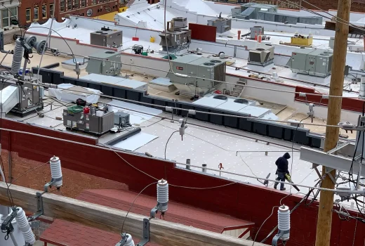 The Keys to Maintaining a Commercial Roof. Detailed view of a commercial rooftop with HVAC units, electrical wiring, and a worker conducting maintenance tasks.