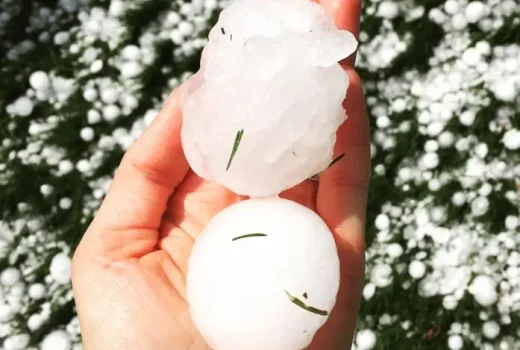 Why Fixing Hail Damage to Your Roof is So Important. Two large hailstones being held in a person’s hand, with a background of hail-covered grass.