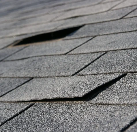 Be Sure To Make Roof Repairs Sooner Rather Than Later. A detailed view of asphalt roof shingles showing signs of damage and lifting, requiring urgent repair.