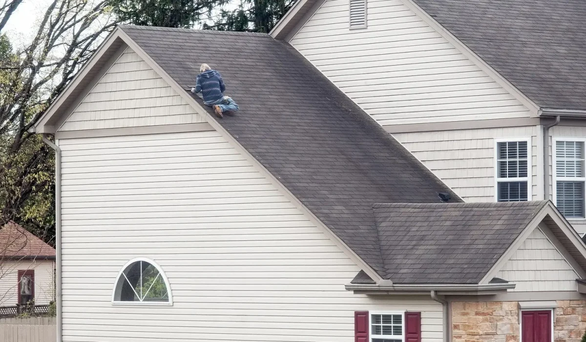 Top Reasons to Go With a Local Home Restoration Company. A roofing professional working on the steep roof of a house, surrounded by tall trees and a cloudy sky.