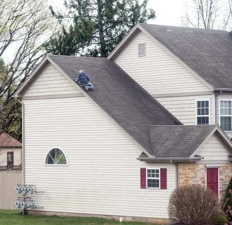 Top Reasons to Go With a Local Home Restoration Company. A roofing professional working on the steep roof of a house, surrounded by tall trees and a cloudy sky.